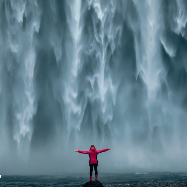 Iceland landscape photo of brave girl who proudly standing with his arms raised in front of water wall of mighty waterfall.