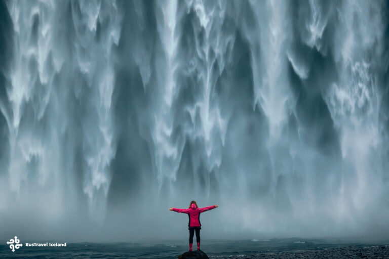 Famous powerful Skogafoss waterfall at south Iceland Iceland landscape photo of brave girl who proudly standing with his arms raised in front of water wall of mighty waterfall.